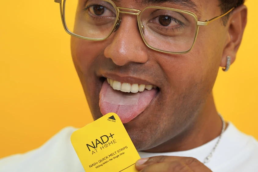 Close-up of a man in glasses with his tongue out, holding a NAD+ oral strip sachet